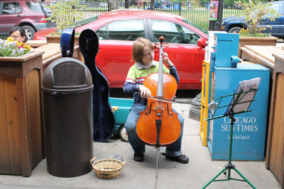 Young teenage busker seated playing cello on a city sidewalk.  Basket of money in front.